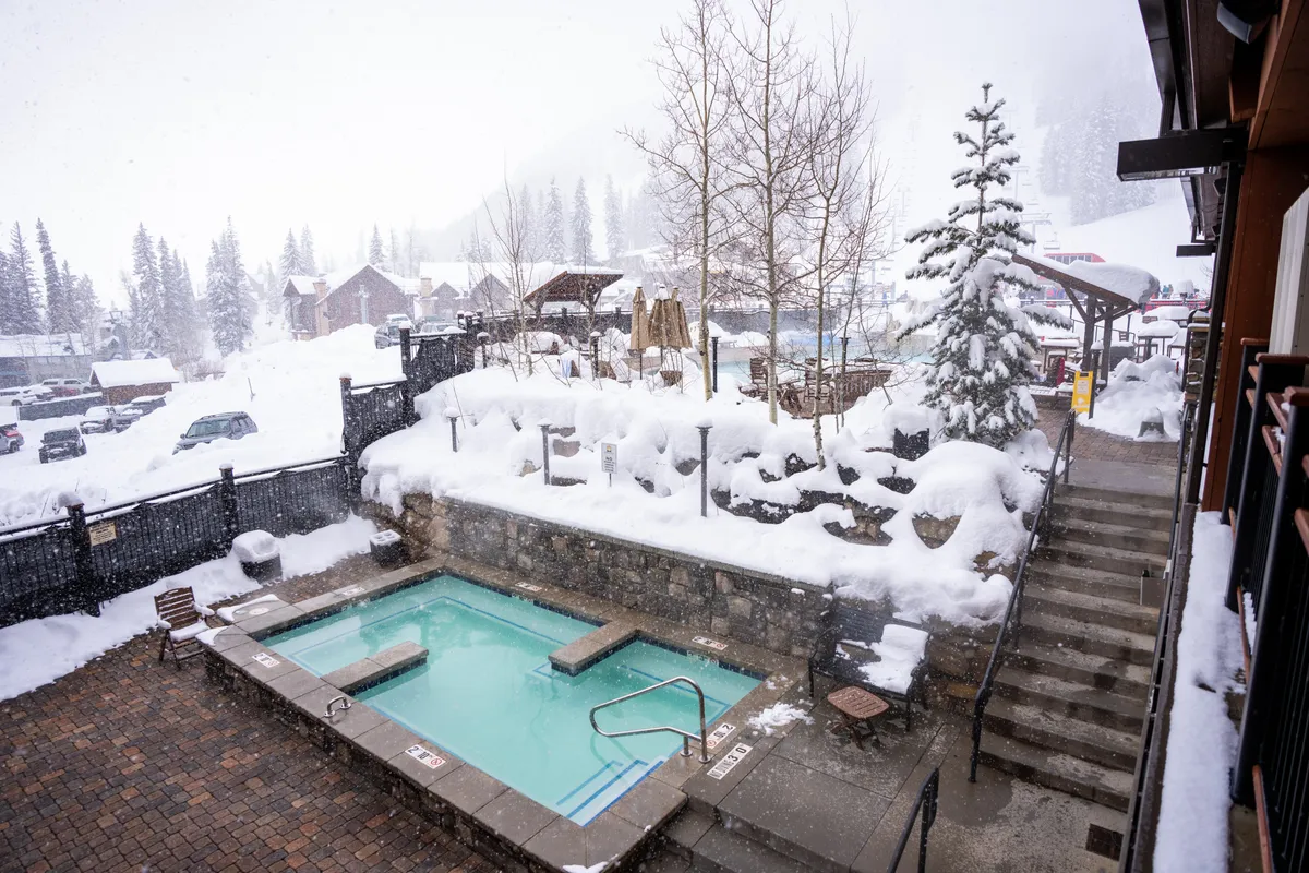 An inviting outdoor hot tub surrounded by a snowy landscape, with steps leading down and tall trees framing the scene. The atmosphere is tranquil and cozy, perfect for relaxing after a day on the slopes.