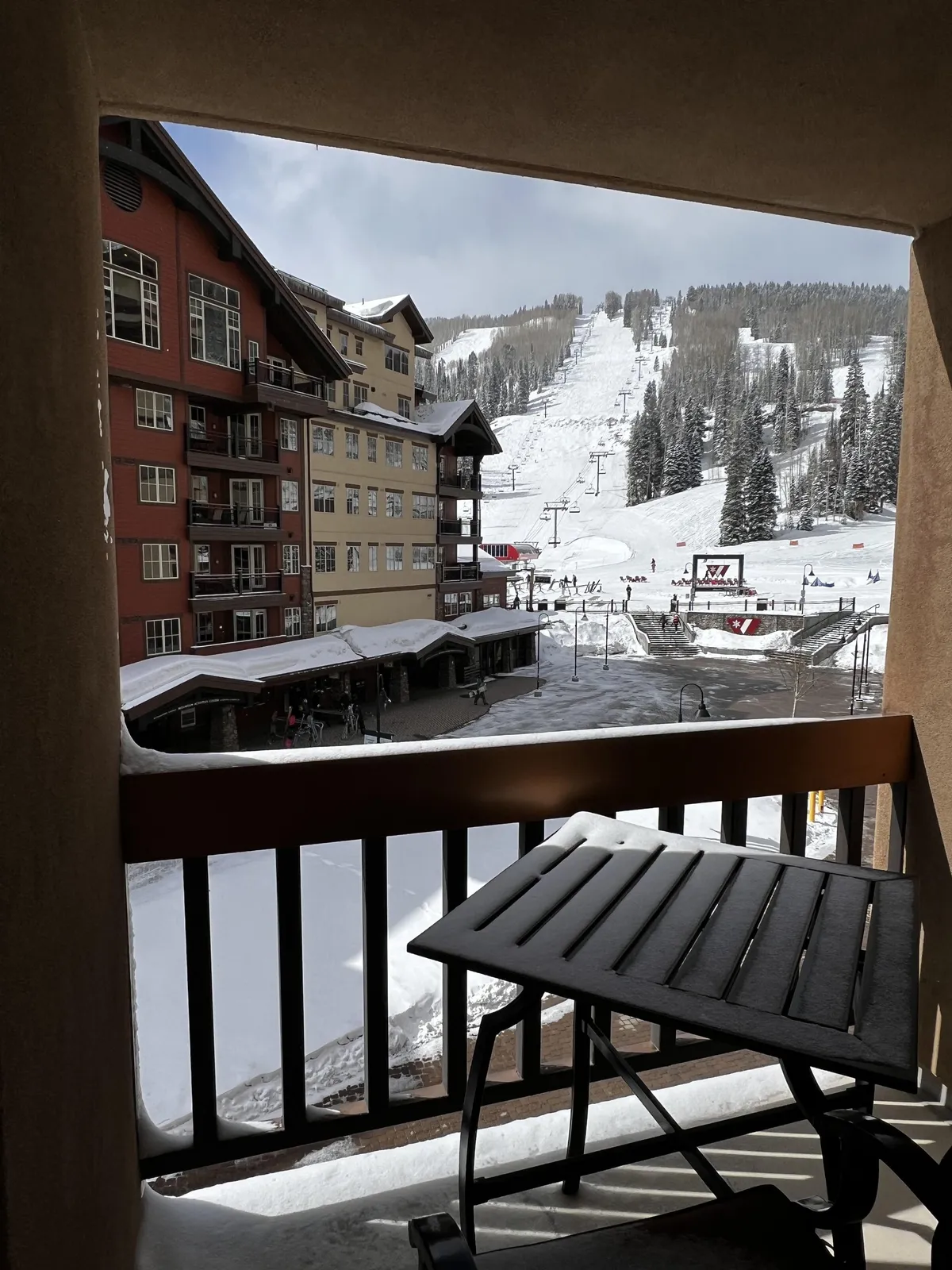 A scenic view from a balcony overlooks the ski slopes, framed by the resort's buildings and snow-covered ground. The image features balcony seating, inviting guests to relax while soaking in the winter scenery and vibrant resort life below.