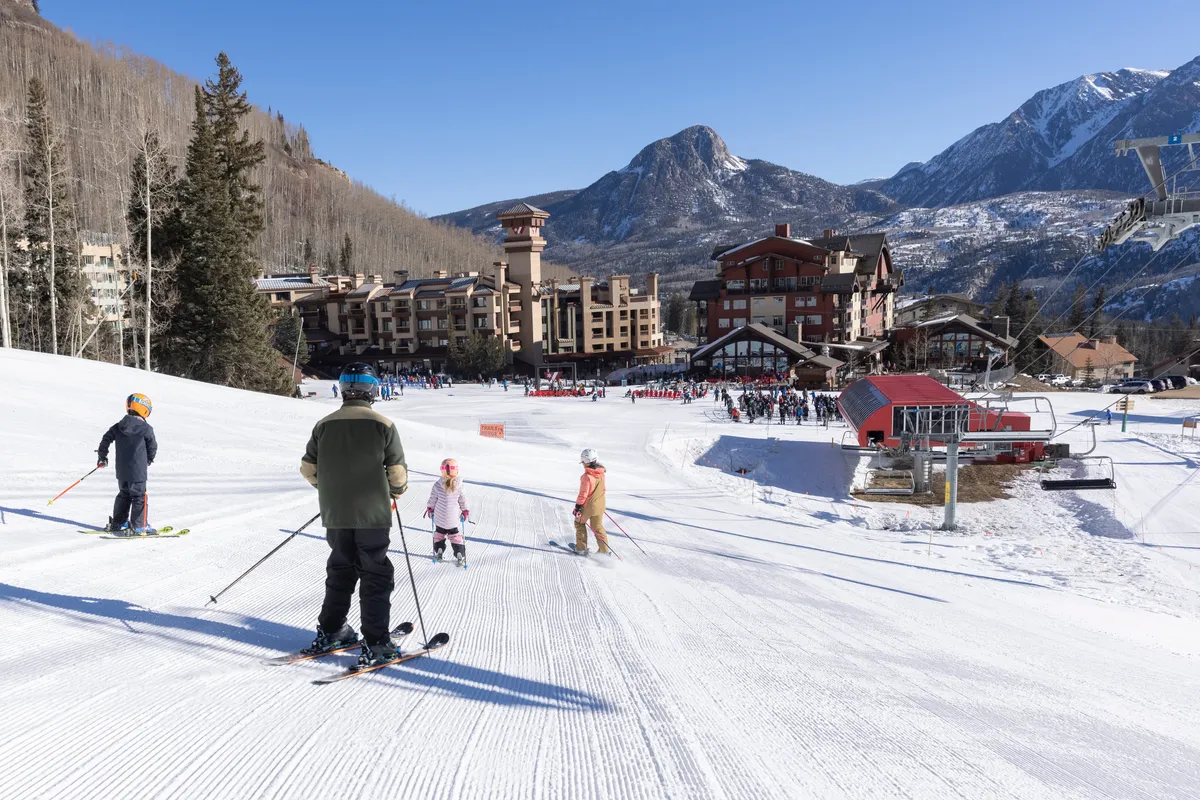 A vibrant winter scene featuring families and skiers enjoying the snowy slopes, with the stunning mountain backdrop and resort buildings visible. The ski lift adds to the atmosphere of fun and adventure, capturing the energy of a mountain vacation.