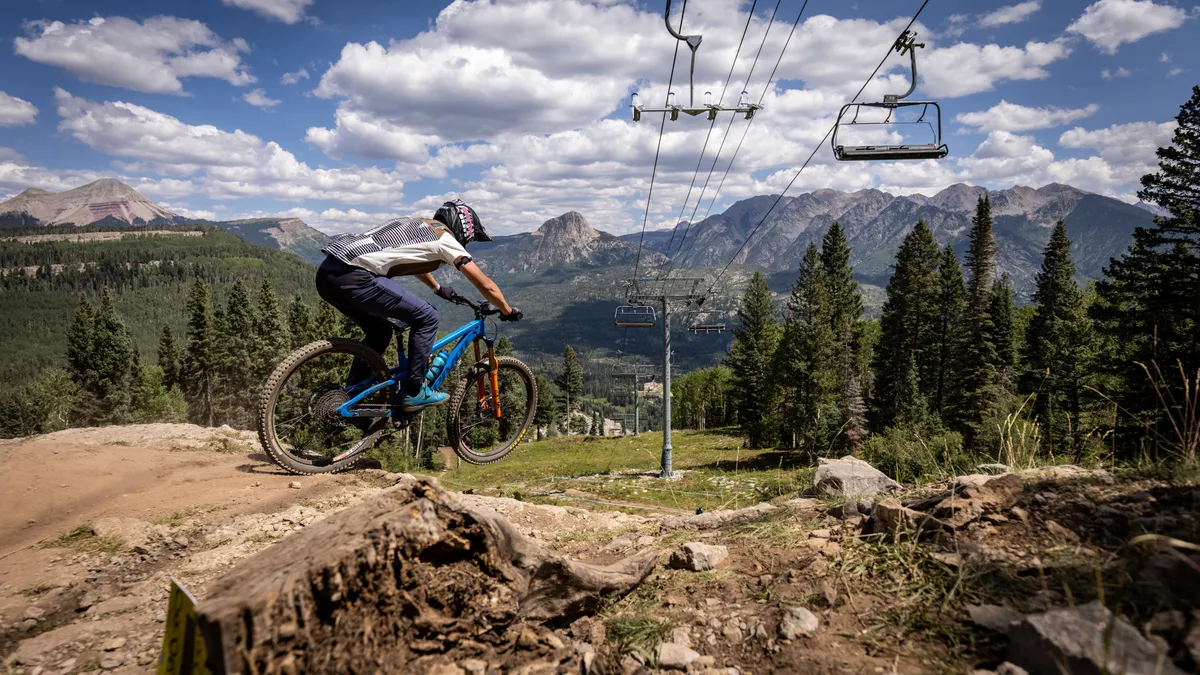 A dynamic exterior shot showing a mountain biker in action against breathtaking mountain landscapes, with a ski lift in the background. The image captures the thrill of outdoor activities like biking, hiking, and skiing.