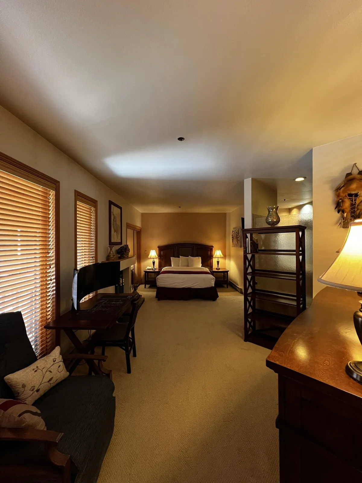 This image features a serene bedroom with a bed dressed in crisp white linens, wooden furniture, a cozy sofa adorned with decorative pillows, and a desk with a chair. Soft lighting from lamps creates a peaceful, inviting atmosphere, perfect for moments of rest and quiet relaxation.