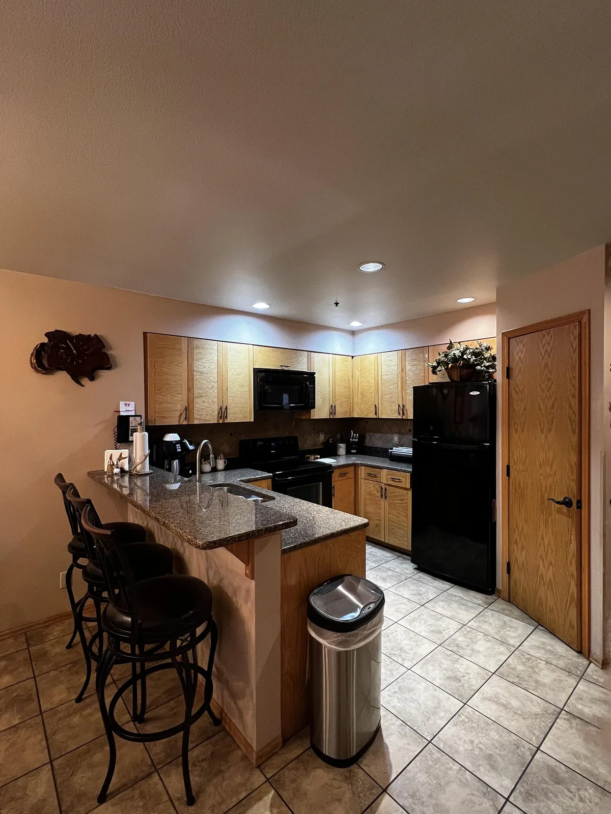 This image highlights the main kitchen area, featuring a bar counter with stools, granite countertop, black appliances, and wooden cabinets. The cozy interior suggests a space designed for both cooking and casual dining, with room for guests or family to gather around the counter.
