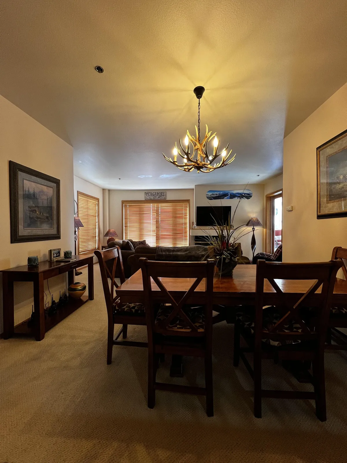 A warmly lit interior featuring a dining table set beneath a classic chandelier, with a plush sofa nearby and decorative plants adding a touch of greenery. The open arrangement suggests a seamless flow between the living and dining spaces, perfect for gatherings and cozy evenings. The atmosphere is inviting, emphasizing comfort and togetherness.
