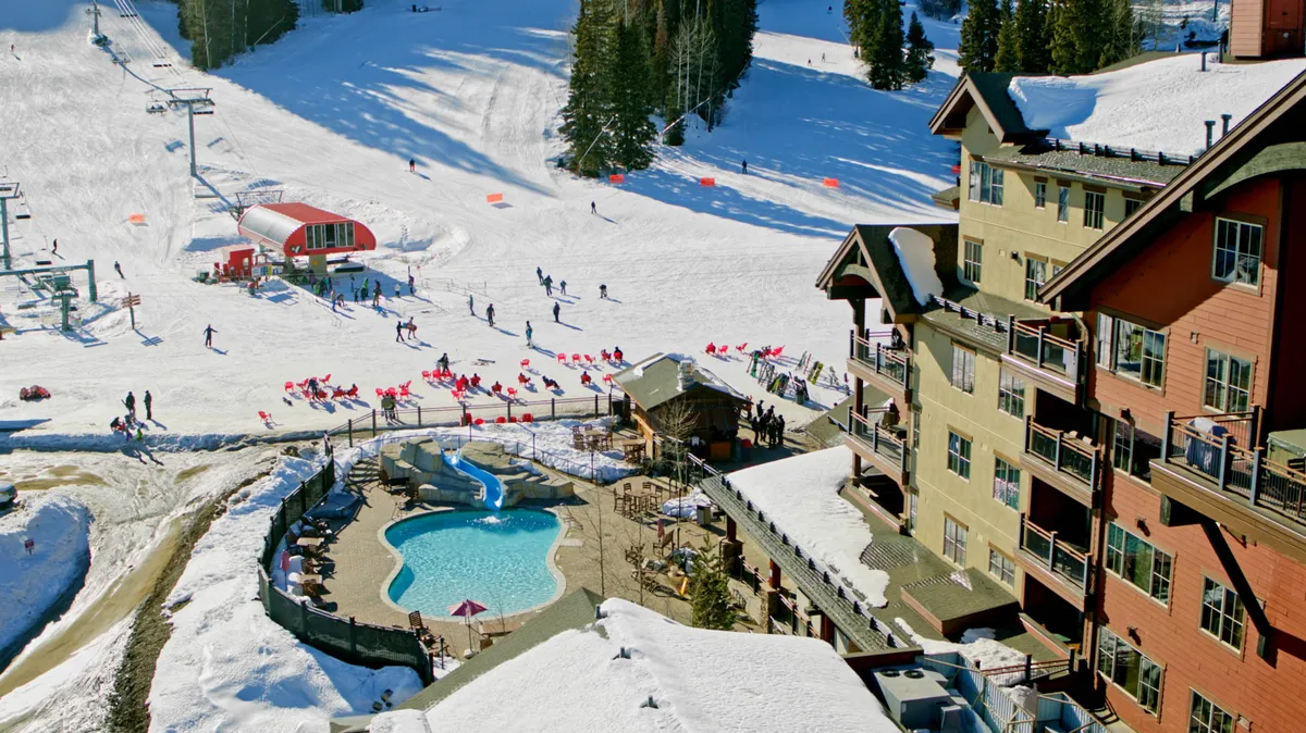 This exterior shot captures the vibrant atmosphere at the base of Purgatory Resort, showing ski slopes, a bustling swimming pool, lodge buildings, and skiers and snowboarders actively enjoying the scene. The snowy landscape, visible resort amenities, and lively activity reflect the energy and accessibility promised in the section.