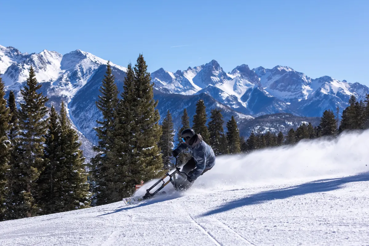 An action shot of a skier carving through pristine snow amidst snow-covered mountains and pine trees. The dynamic motion and dramatic mountain backdrop evoke the thrill and adventure of mountain sports.