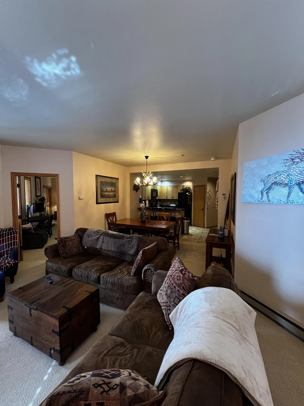 A spacious living area featuring a large brown sectional sofa and a wooden coffee table, with the dining area and chandelier visible in the background. Wall art and warm lighting contribute to a cozy, mountain-lodge vibe. The open layout shows how the living and dining spaces connect.